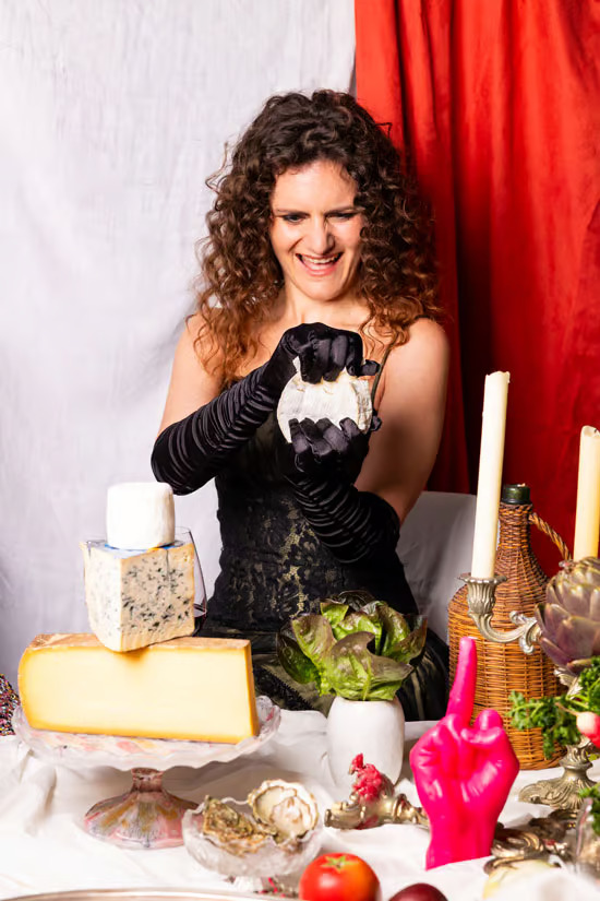 Emily Monaco in black opera gloves holding a knife over a spread of cheeses against a red curtain