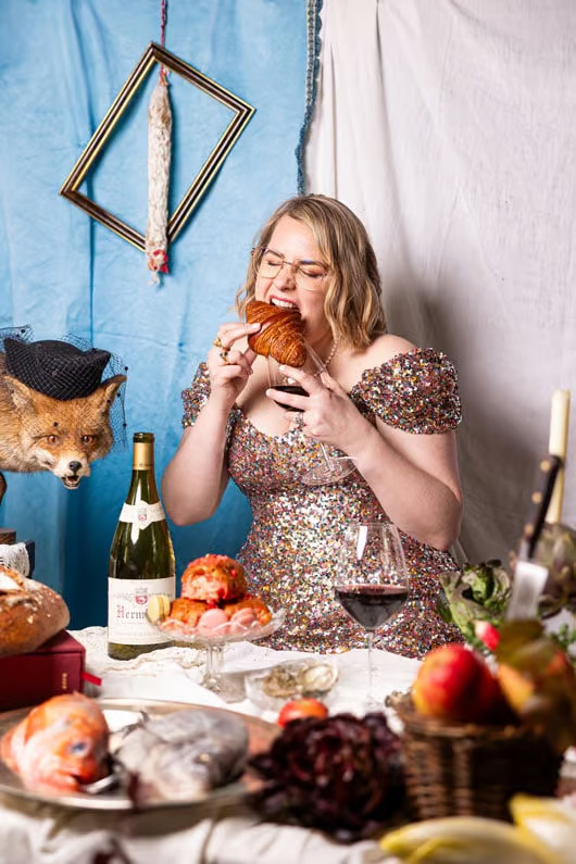 Caroline Fazeli in a sequined dress biting into a croissant at a candlelit table set with wine and food