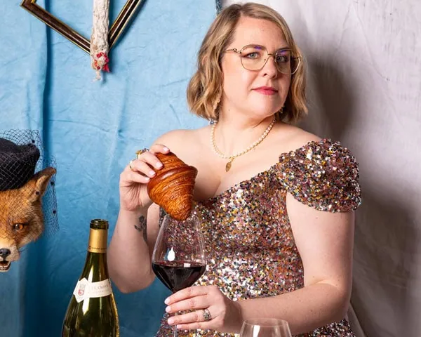 Caroline Fazeli in a sequined dress holding a croissant beside a bottle of white wine
