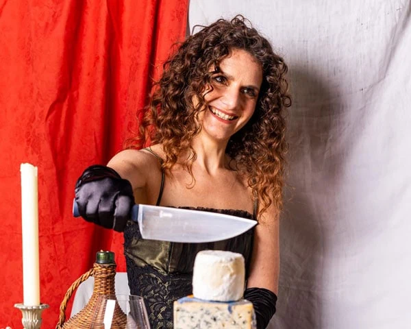 Emily Monaco slicing a wheel of cheese in front of a red curtain