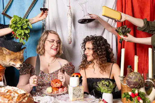 Caroline and Emily toasting at a candle-lit table framed by red curtains